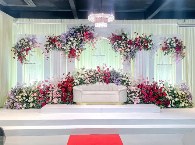 White bridal sofa surrounded by pink, red, white and lilac flowers, illuminated columns and a chandelier in a past Joy Events & Catering stage setup