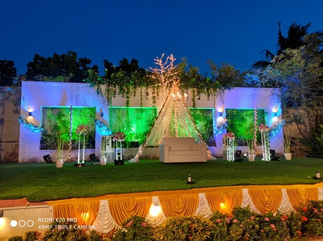 Outdoor lawn stage with a white sofa, triangular sheer canopy, fairy lights, flower stands and blue and green event lighting from Joy Events & Catering's past décor work
