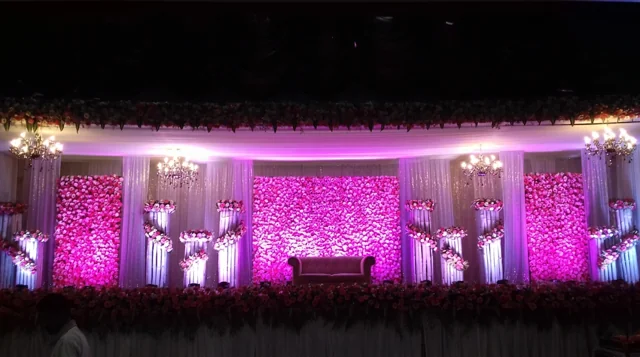 Wide reception stage illuminated in magenta with three flower panels, hanging chandeliers, tiered floral stands and central bridal seating from Joy Events & Catering's past décor