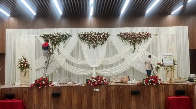 Two décor team members arranging flowers and ivory fabric swags, one on a ladder, during preparation of a past Joy Events & Catering stage