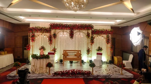 Gold arch frames topped with red and white flowers, chandeliers and fairy lights behind a wooden bridal bench at a past Joy Events & Catering event