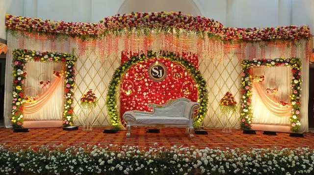 Deep-red rose floral wall with a circular monogram emblem, white carved chaise sofa, ivory draped backdrop with gold lattice frames and hanging pink garlands from Joy Events & Catering's past décor