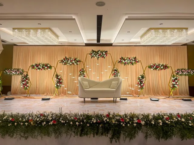 Hexagonal gold frames holding red, pink and white flower arrangements in front of peach pleated drapes, a curved cream sofa, crystal chandeliers and a greenery flower bed from Joy Events & Catering's past décor